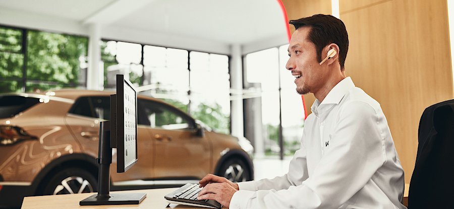 Ein Mann im weißen Hemd arbeitet am Computer in einem modernen Autohaus mit einem Auto im Hintergrund.