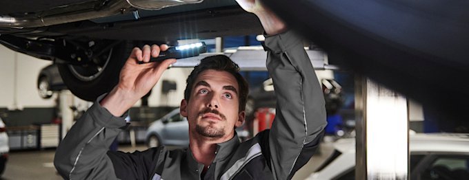 Mechaniker arbeitet unter einem Auto mit Taschenlampe in einer Werkstatt.