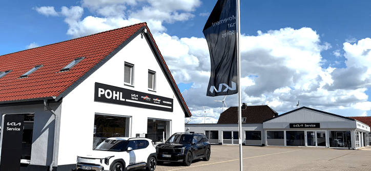 Autohaus Pohl mit zwei Autos und einem Flaggenmast vor einem blauen Himmel.